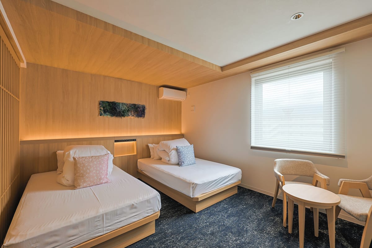 A hotel room with two beds, a small table, and a window with blinds.