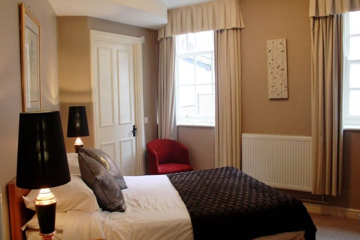 Una habitación de hotel con cama, una silla roja, una lámpara y una ventana con cortinas.