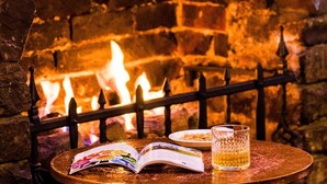 Une table ronde en bois, sur laquelle est posé un magazine ouvert, un verre contenant un liquide ambré et une assiette de nourriture. Au fond, on aperçoit un mur de briques avec une cheminée.