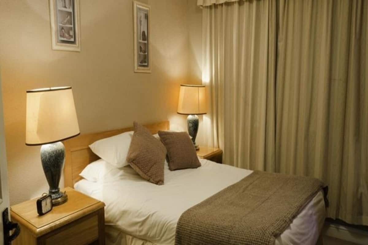 Una habitación de hotel con cama, dos lámparas de noche, una mesita de noche con reloj y cortinas.