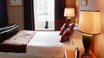 Una habitación de hotel con cama, mesita de noche con lámpara, ventana con cortinas y vistas a edificios.