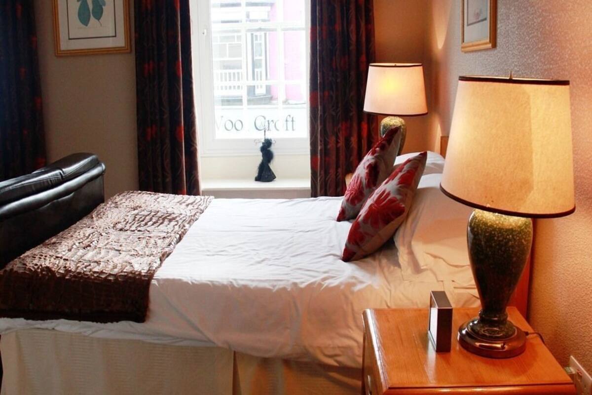 Habitación de hotel con cama, dos lámparas de noche, mesita auxiliar y vista a un edificio a través de una ventana.
