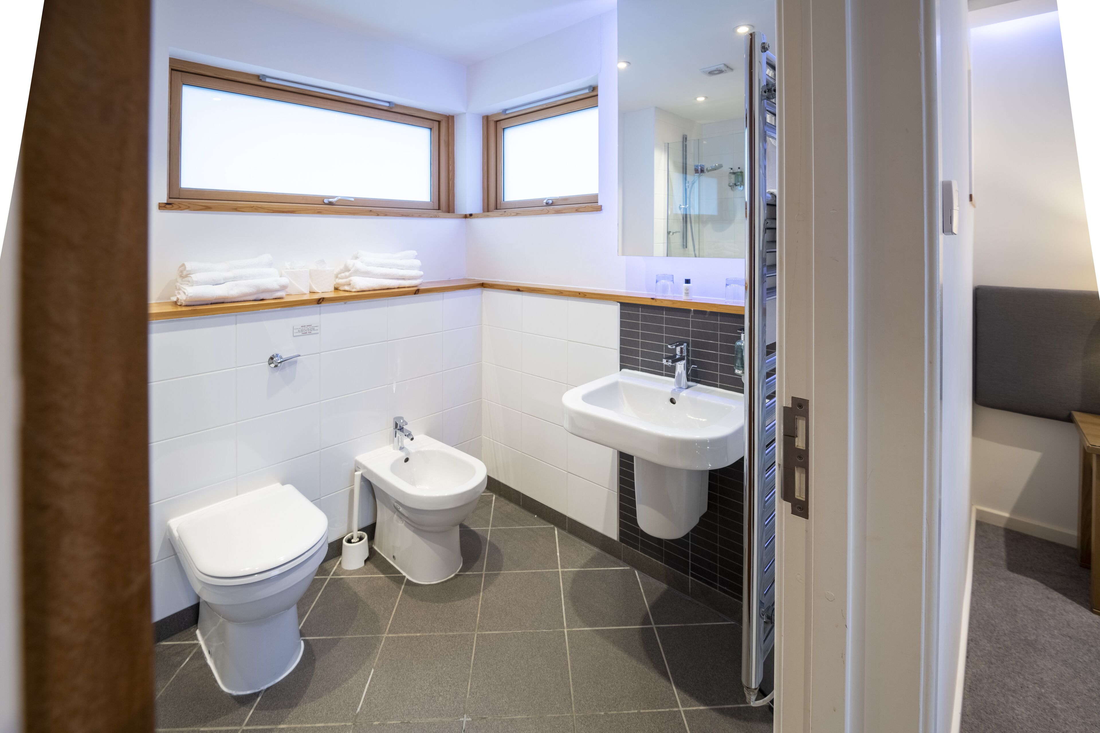 A modern bathroom with a toilet, bidet, and sink. There are two windows above the toilet and a shower area visible.