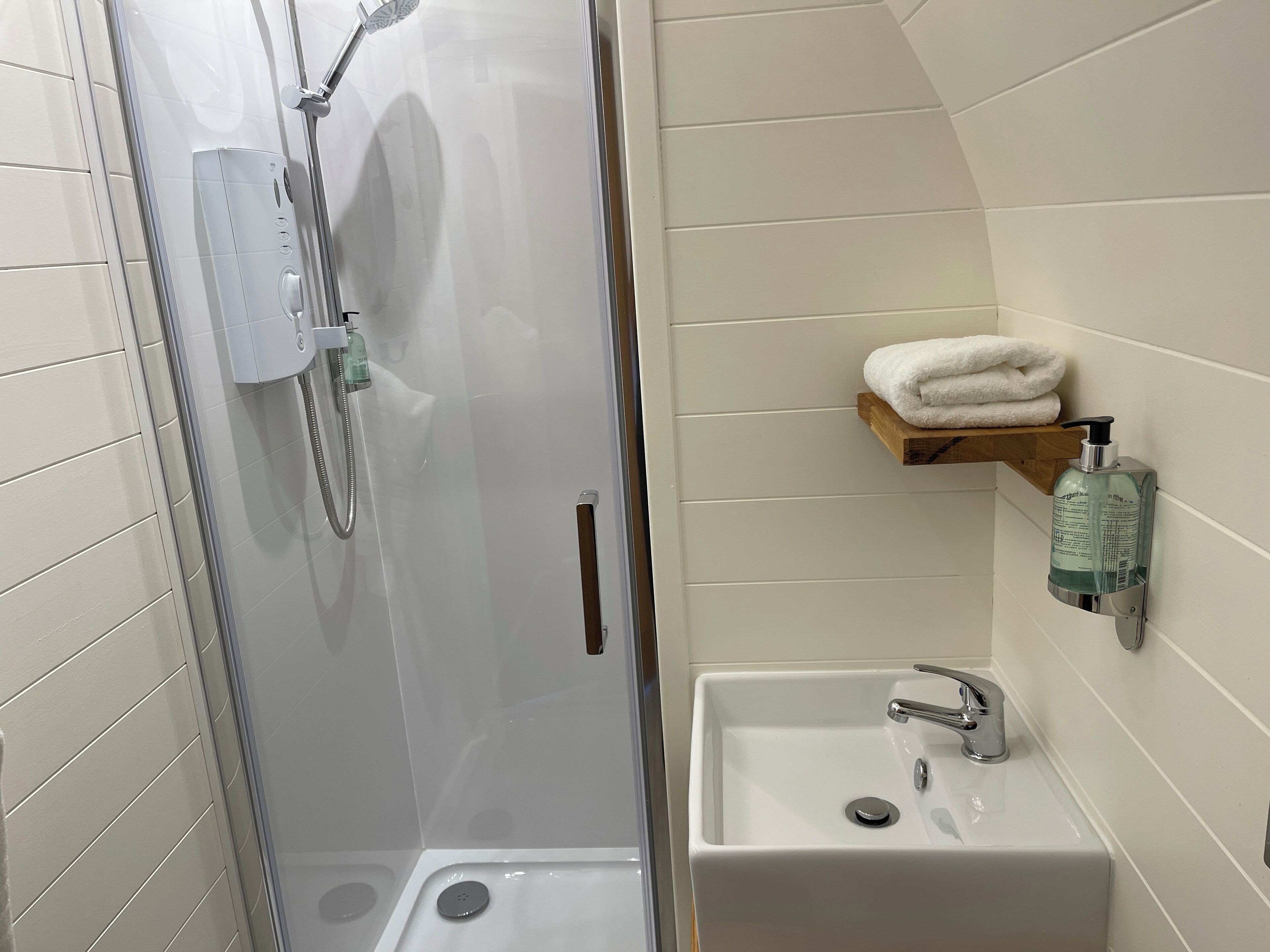 A bathroom with a glass-enclosed shower, a sink with a faucet, and a shelf with a towel and a soap dispenser.