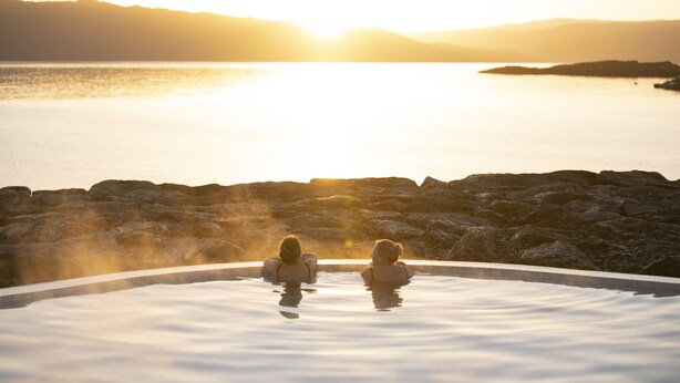 Two people in a hot tub with a sunset view over water and mountains.