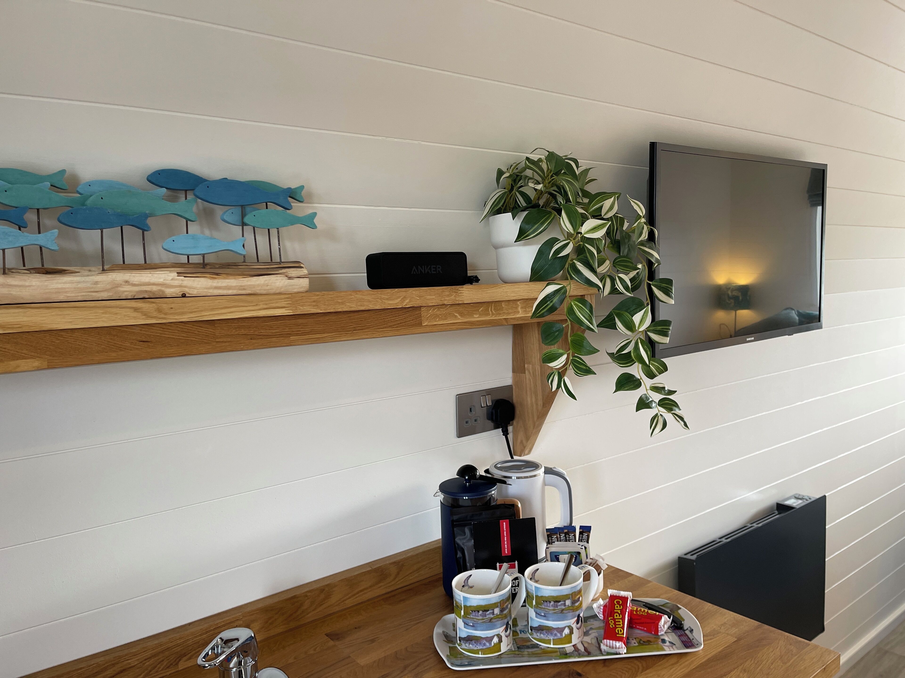 Une table en bois, avec une bouilloire, deux tasses, un plateau de collations et une plante posée sur une étagère au-dessus.