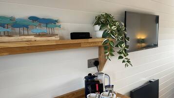 A wooden table with a kettle, two mugs, a tray with snacks, and a plant on a shelf above.