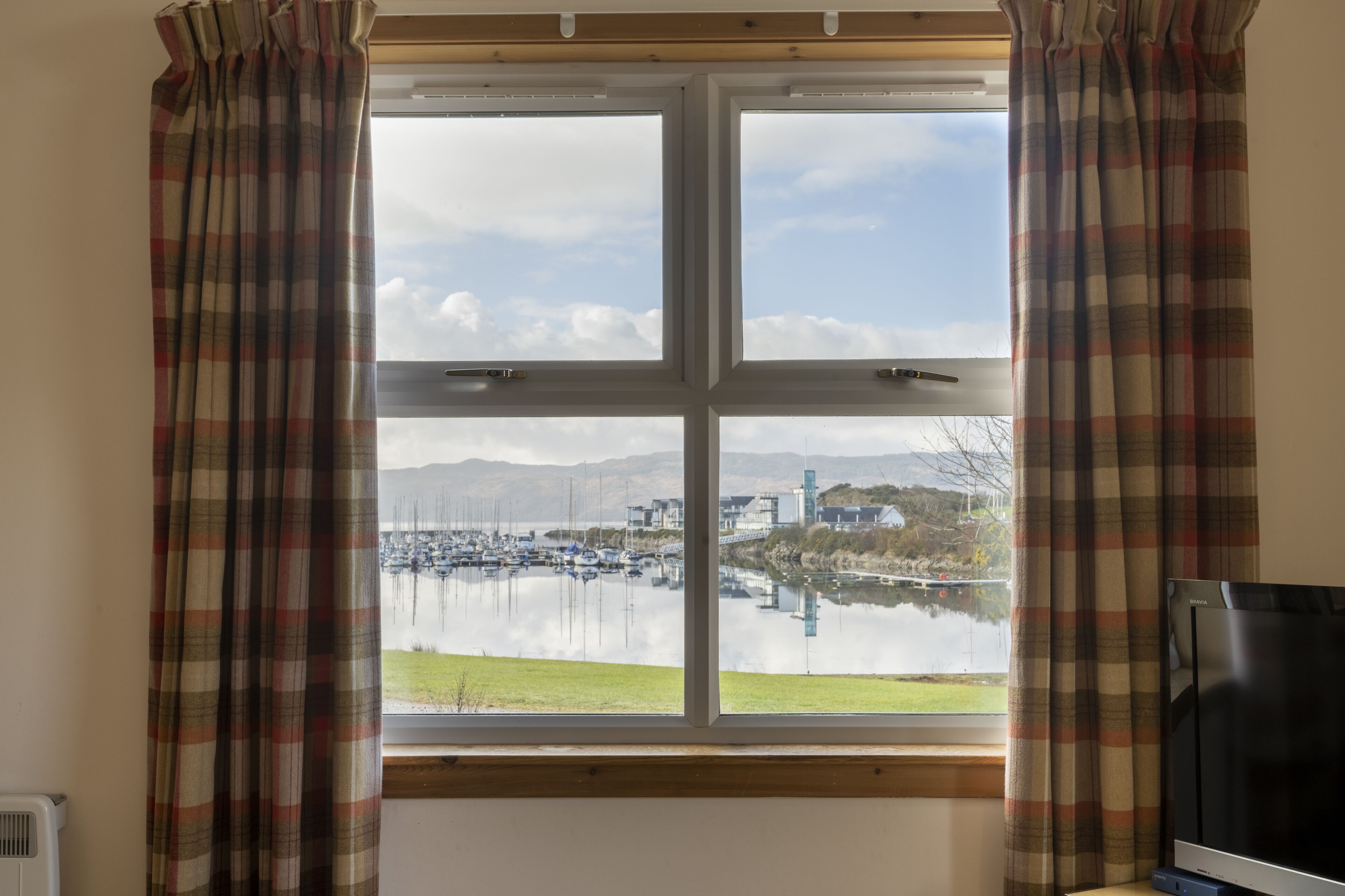 Una ventana con cortinas a cuadros, vista de un puerto deportivo con barcos y un edificio al otro lado del agua.