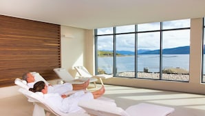 A spa room with reclining chairs, a wooden wall, and a large window overlooking a lake and mountains.
