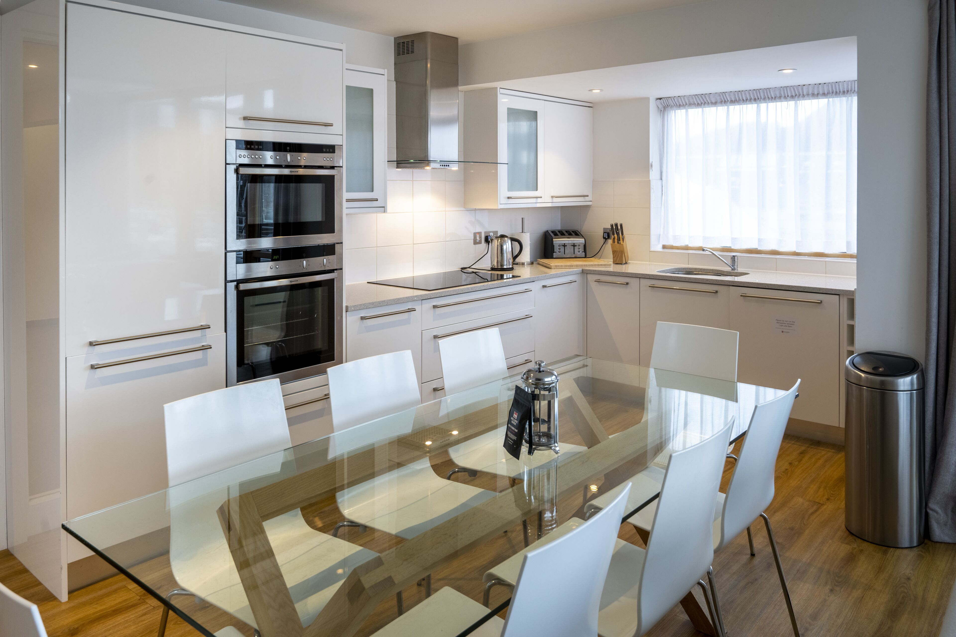 A modern kitchen with white cabinetry, stainless steel appliances, a glass dining table, and a window with curtains.