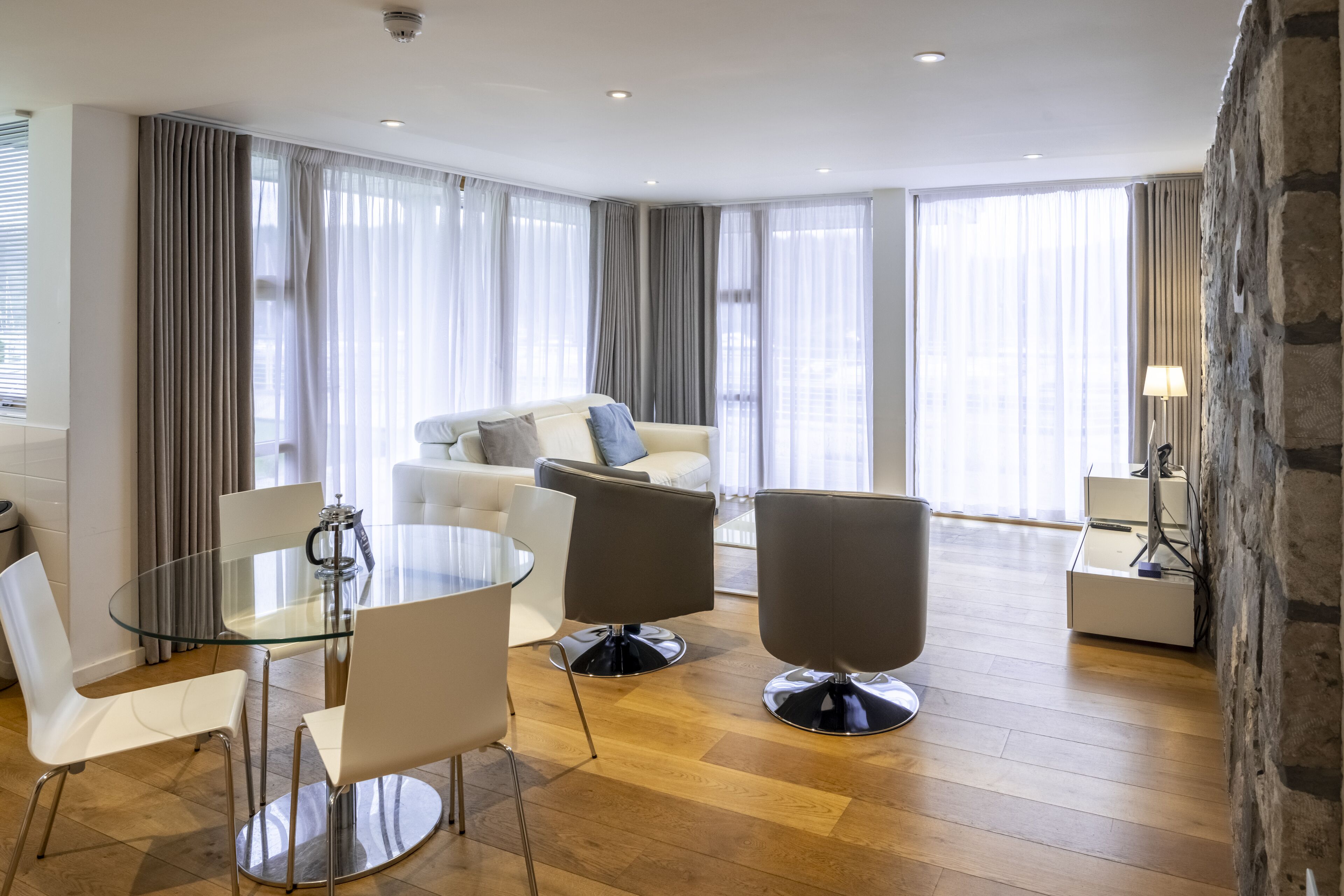 A modern living room with a glass dining table, white and gray chairs, a gray sofa, and a stone accent wall.