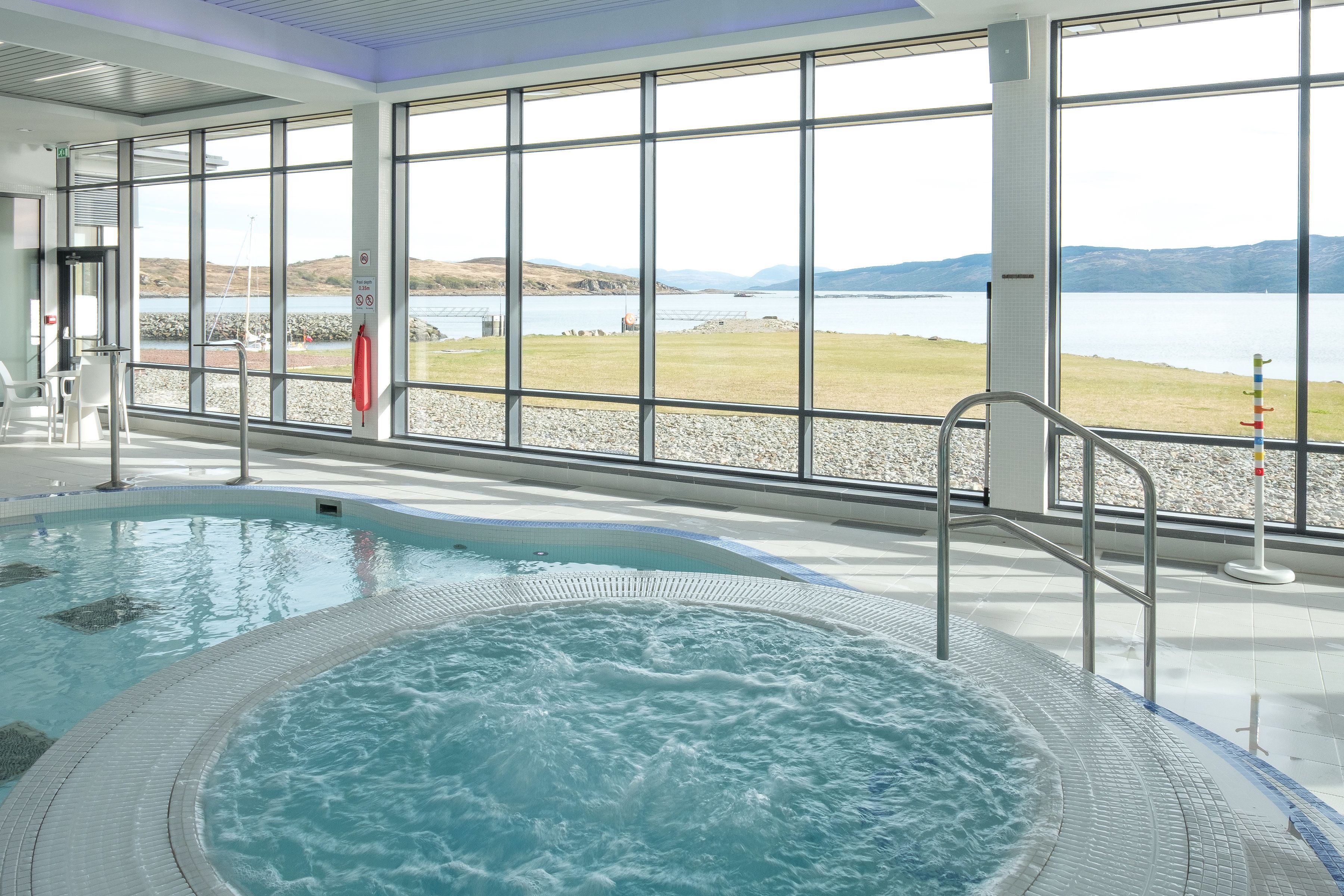 A swimming pool with a hot tub, large windows, and a view of a lake and mountains.