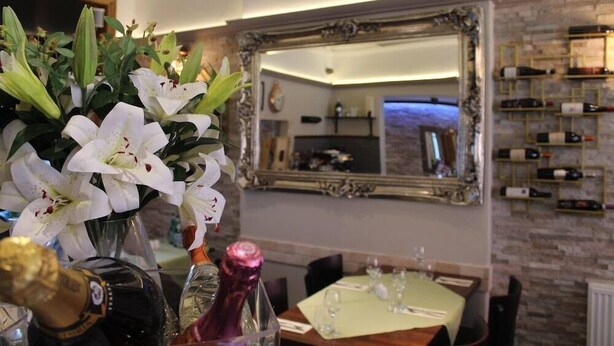 A dining area with a table set for two, a vase of white flowers, and a wine rack with bottles. - Elgano (Cardiff)