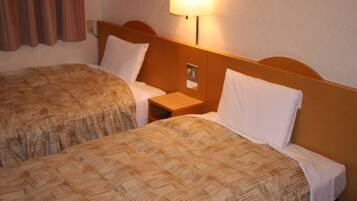 Quarto de hotel com duas camas, cada uma com colcha estampada e almofadas brancas. Há uma mesa de cabeceira entre as camas, com um abajur em cima.