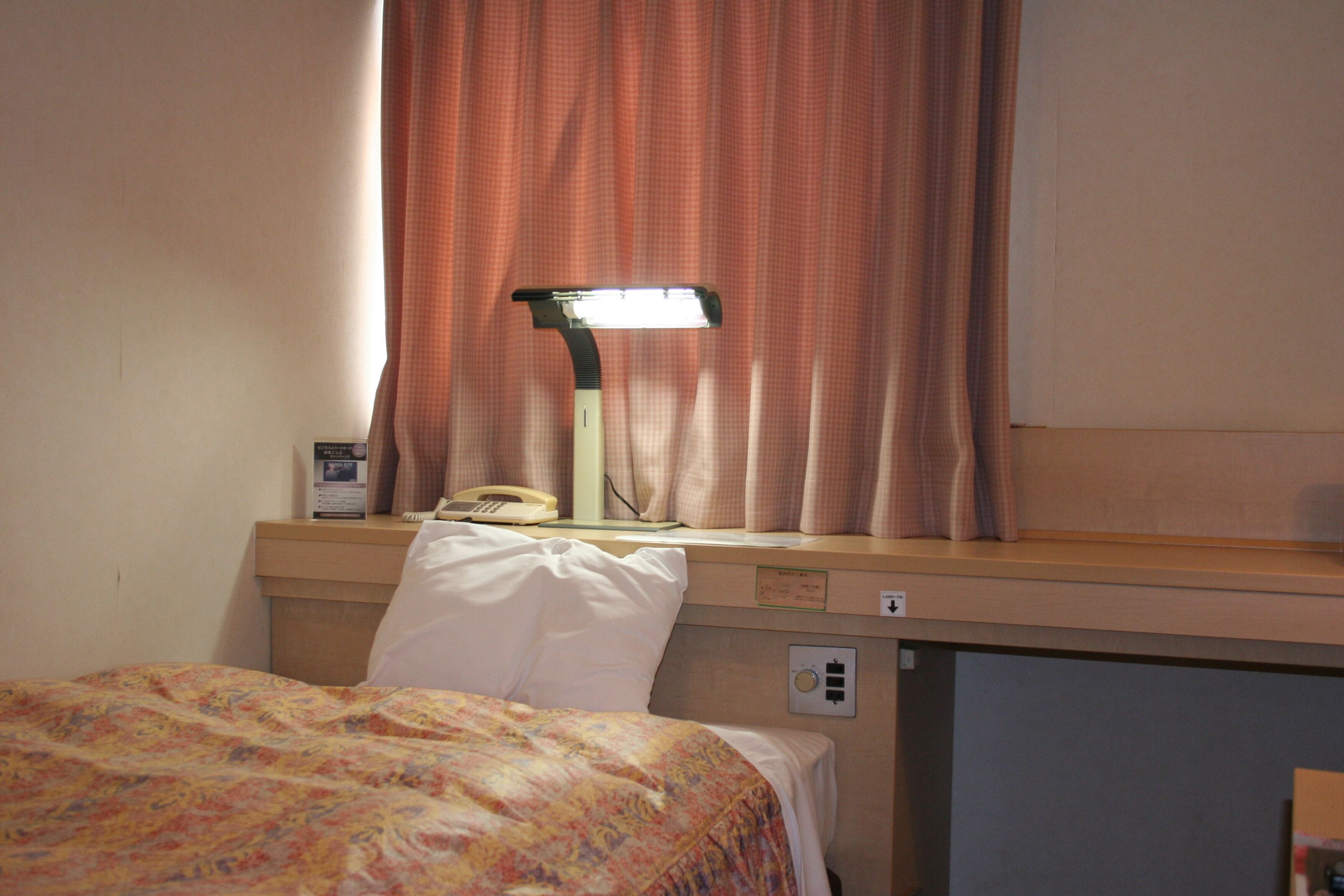Een hotelkamer met een bed, een bureau met een lamp, een telefoon en een raam met gordijnen.