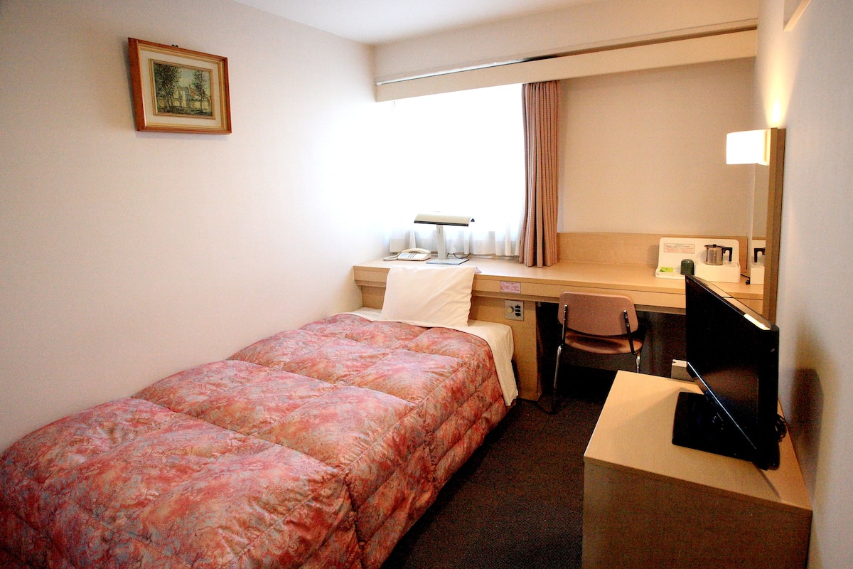 Ein Hotelzimmer mit einem Bett, einem Schreibtisch mit Computer, einem Sessel und einem Fenster mit Vorhängen.