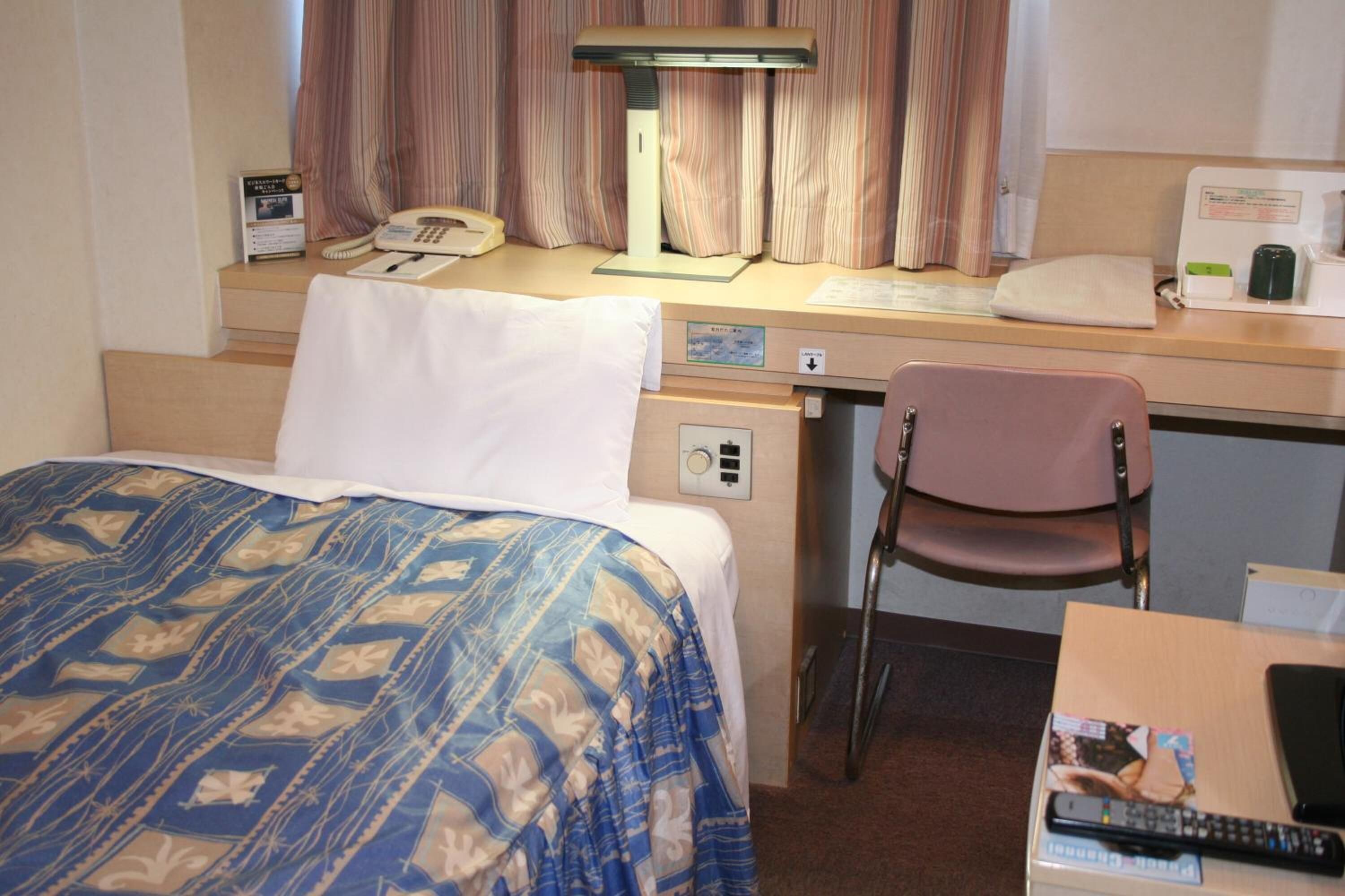 A hotel room with a bed, desk, chair, desk lamp, telephone, and a remote control on a table.