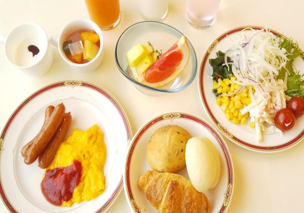 A breakfast table with plates of food including sausages, scrambled eggs, croissants, and a salad with corn, tomatoes, and shredded cheese.