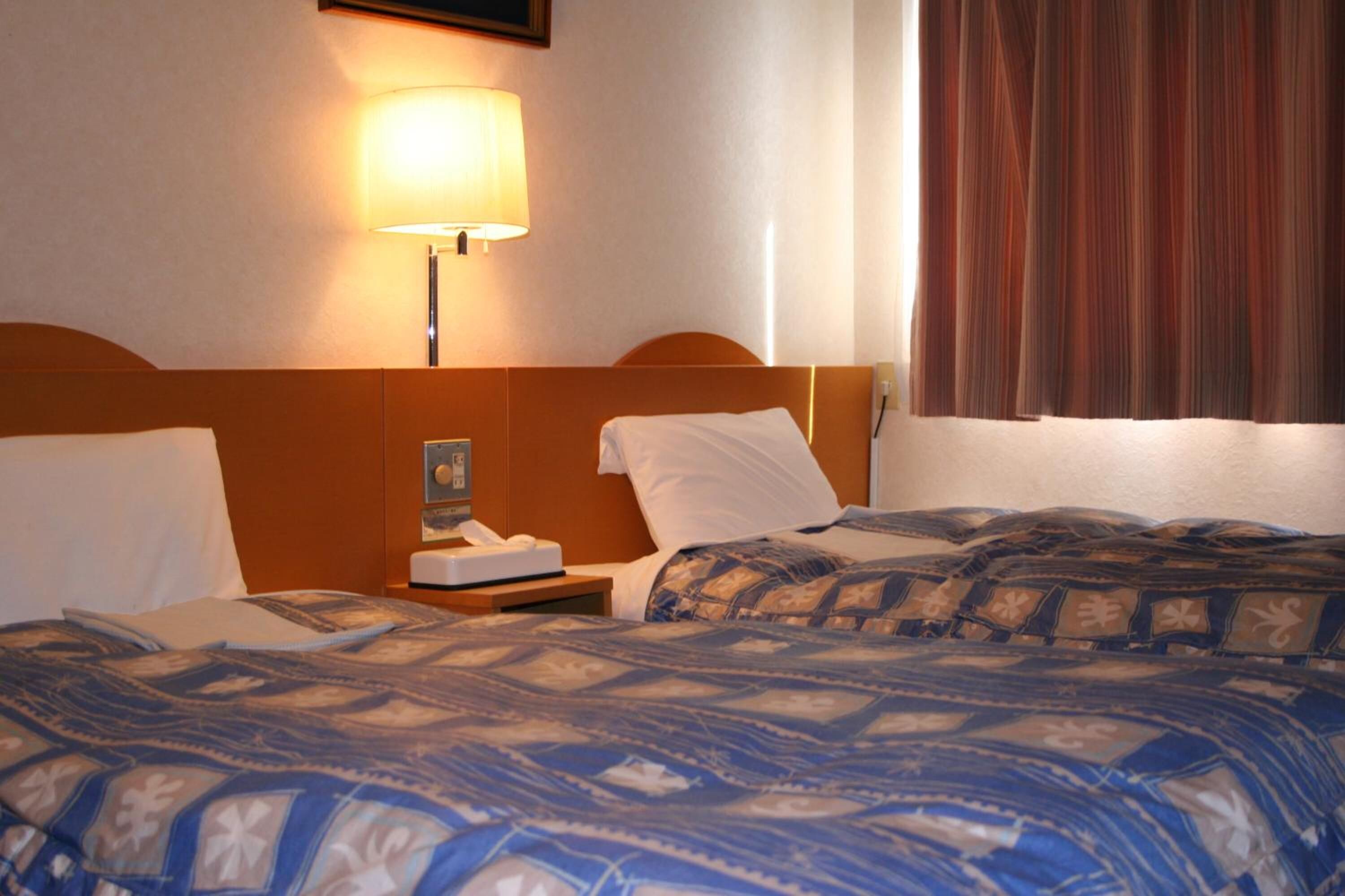 Habitación de hotel con dos camas, una mesita de noche, una lámpara y un ventanal con cortinas.
