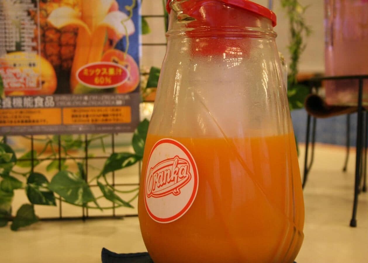 Una botella de vidrio con tapón rojo llena de líquido anaranjado, etiquetada como “Oranka”, sobre una mesa con un fondo que incluye un cartel con imágenes de frutas y texto.