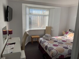 A hotel room with a bed, a desk with a lamp, a chair, a window with blinds, and a TV mounted on the wall.
