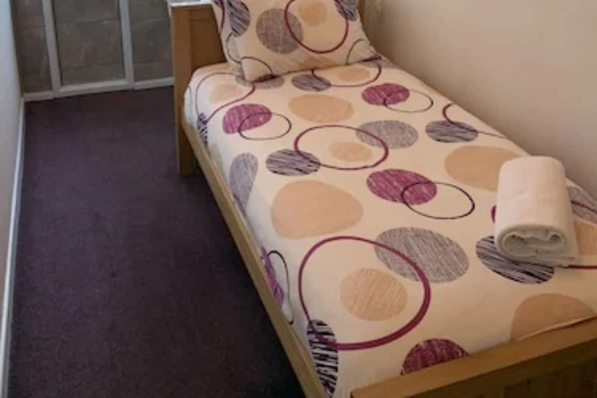 A small, single-bed room with a wooden headboard, a patterned bedspread, a window with blinds, a small kitchenette, and a framed picture on the wall.