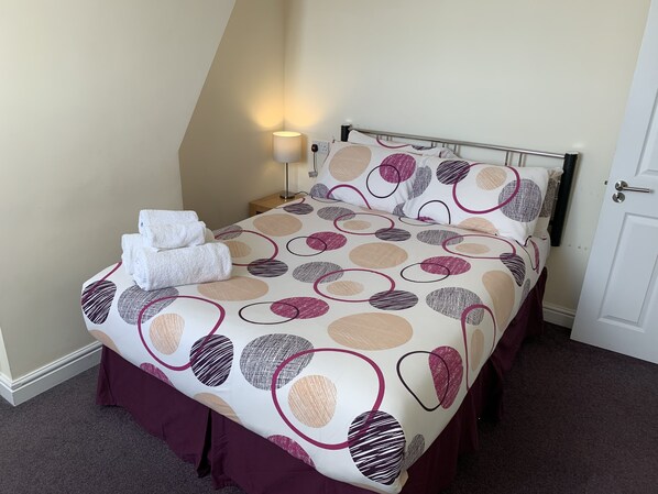 A neatly made bed with a patterned bedspread, a bedside lamp, and a stack of white towels. - Austins Self Service Guest House (Cardiff)