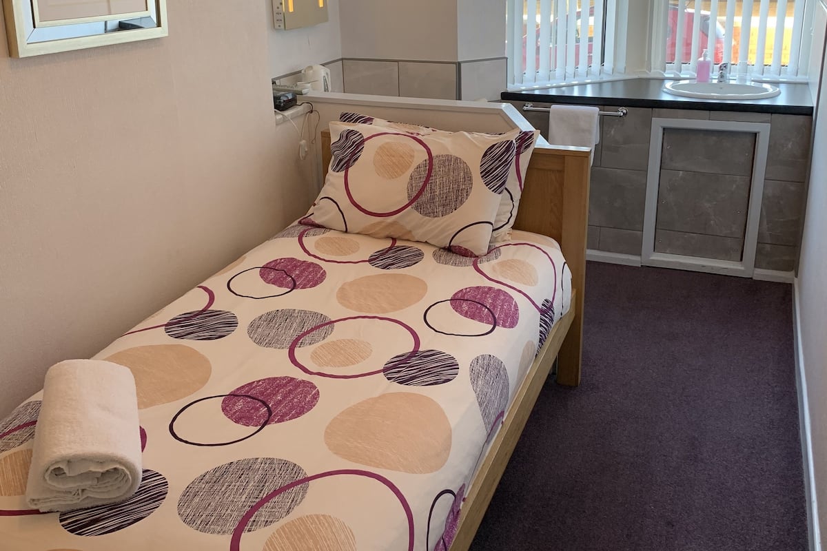 A small, single-bed room with a wooden headboard, a bedside table, a framed picture on the wall, and a window with blinds.