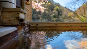 A wooden structure with a water feature, a wooden bench, and a view of cherry blossoms and hills in the background. - Natsuse Onsen Miyakowasure (Senboku)
