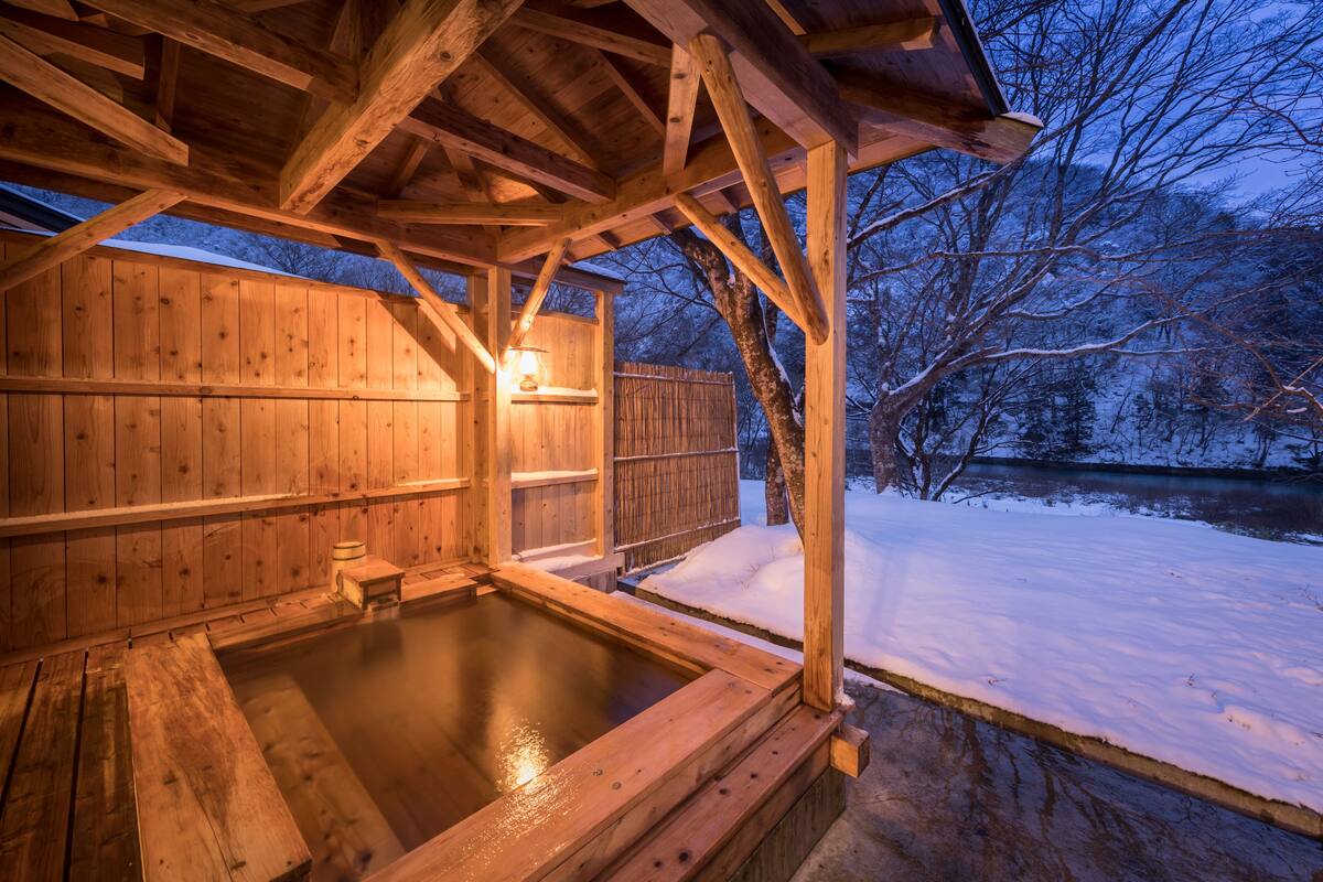 A wooden hot tub under a wooden roof with a view of a snowy landscape.