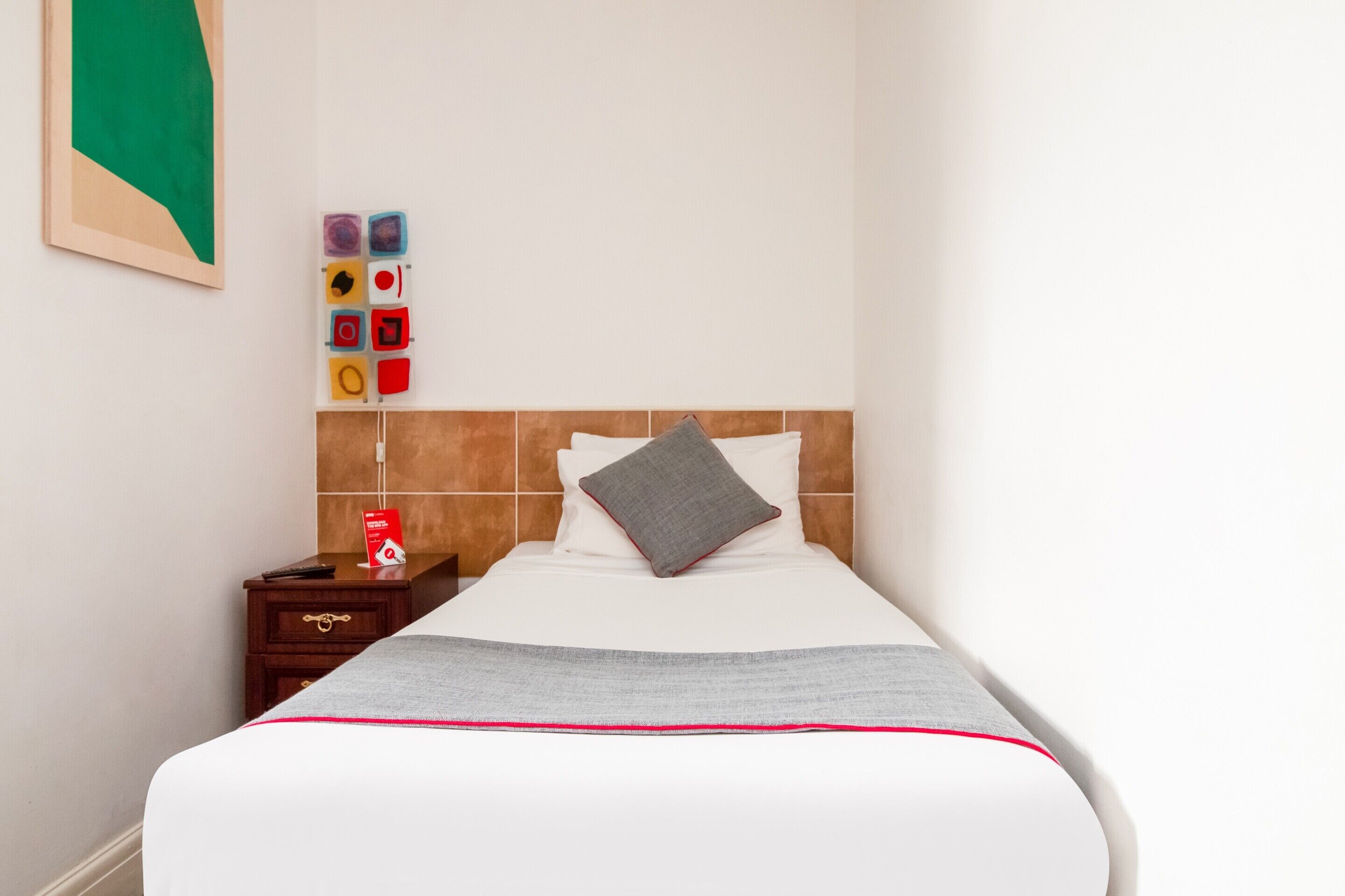 A neatly made bed with a grey and white bedspread, a wooden nightstand with a red object on it, and a wall-mounted light switch.