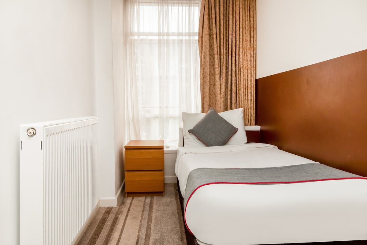 A neatly arranged hotel room with a single bed, a nightstand, a radiator, and a window with sheer curtains.