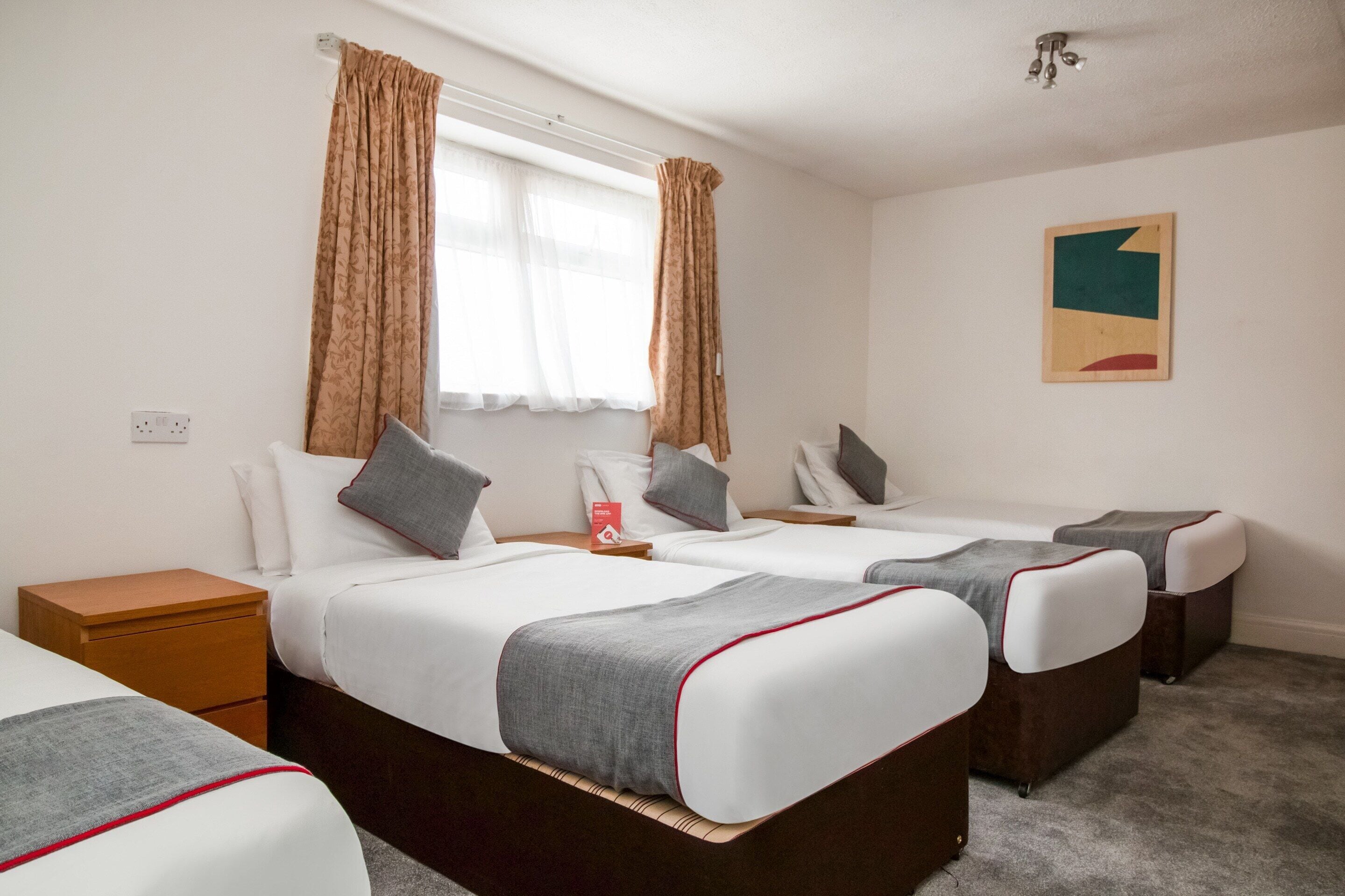 A hotel room with two single beds, a nightstand, a bedside table, a window with curtains, and a wall-mounted artwork.