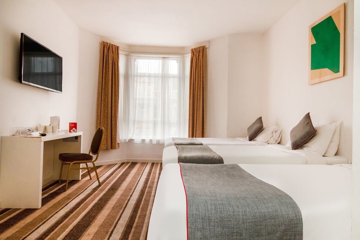 A hotel room with a desk, chair, two beds, a TV, and a window with curtains.