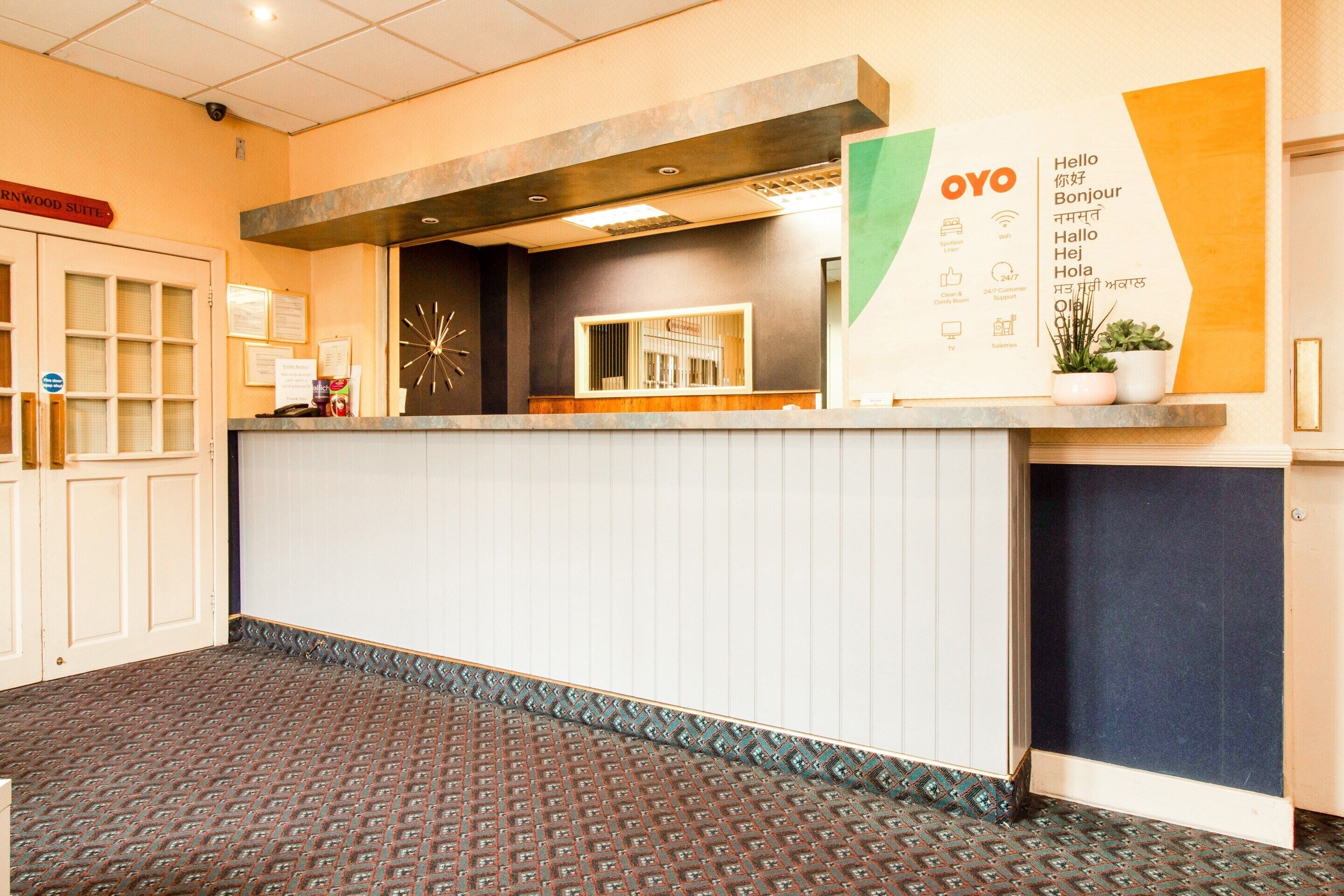 Een receptiezone met een receptiebalie, een klok, een kamerplant en een bord met meertalige begroetingen.