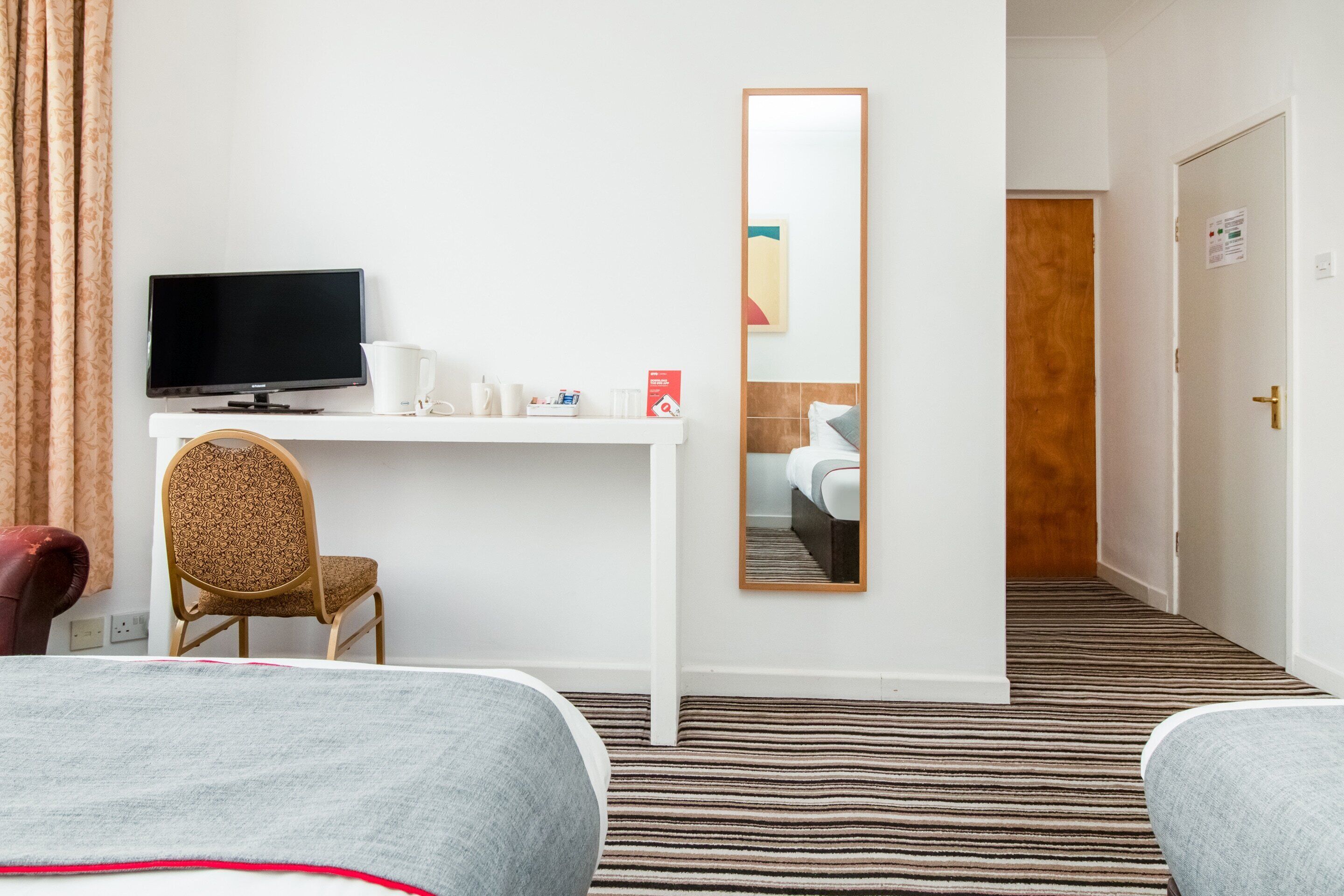 Una habitación de hotel con una cama, un escritorio con computadora, una silla, un espejo y una puerta.