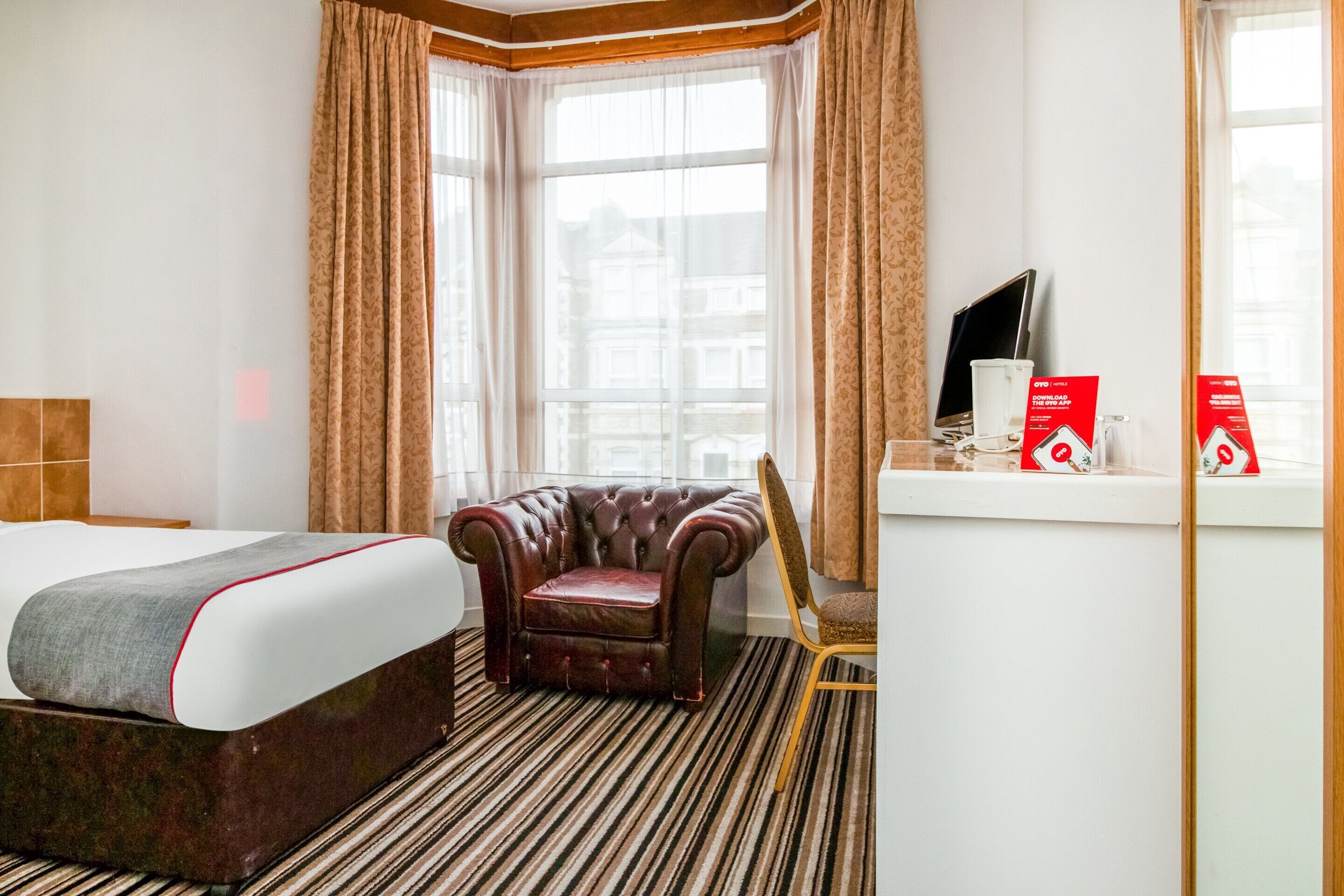 A hotel room with a large bed, a leather armchair, a small table with a TV and red boxes, and a window with curtains.