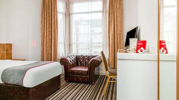 A hotel room with a large bed, a leather armchair, a small table with a TV and red boxes, and a window with curtains.