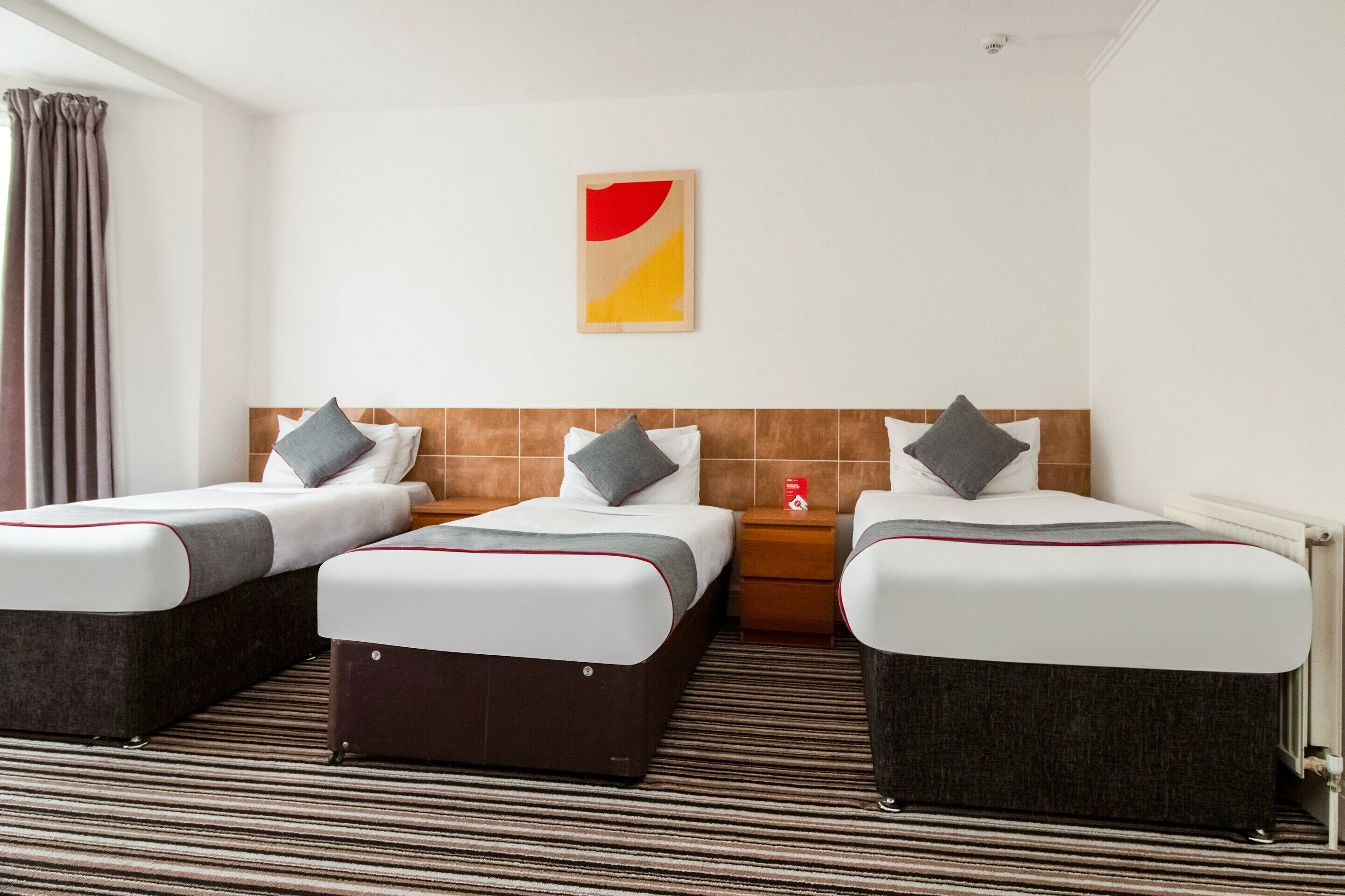 Habitación de hotel con tres camas, cada una con almohada y manta. Se aprecia cabecera de madera, alfombra a rayas y una obra de arte colgada en la pared.