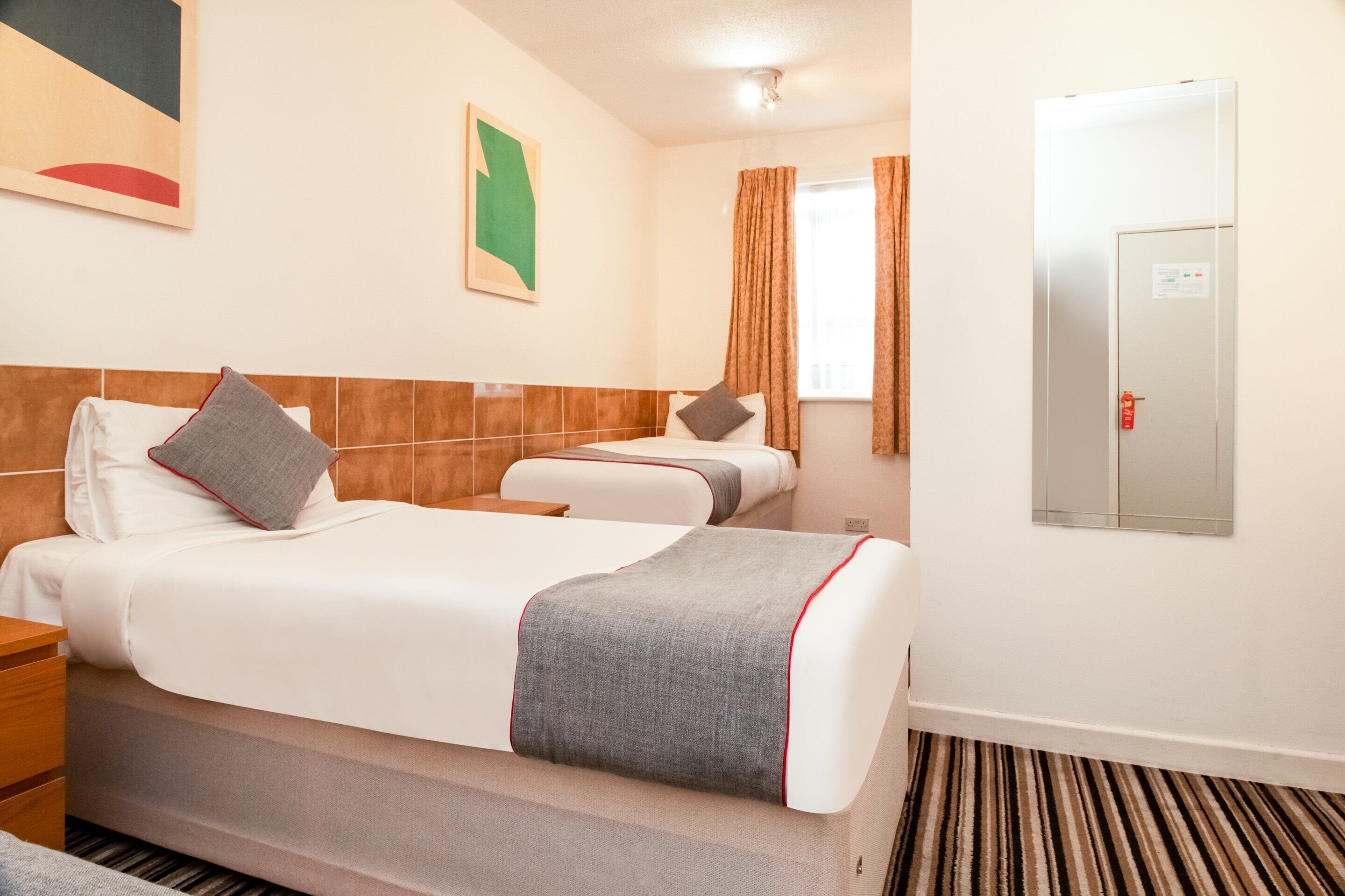 Ein Hotelzimmer mit einem Bett, einem Nachttisch, Vorhängen an einem Fenster und einer Tür mit Rauchmelder.