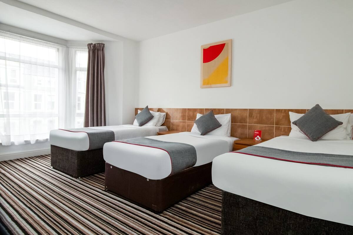 A hotel room with two beds, a window with curtains, a striped carpet, and a painting on the wall.
