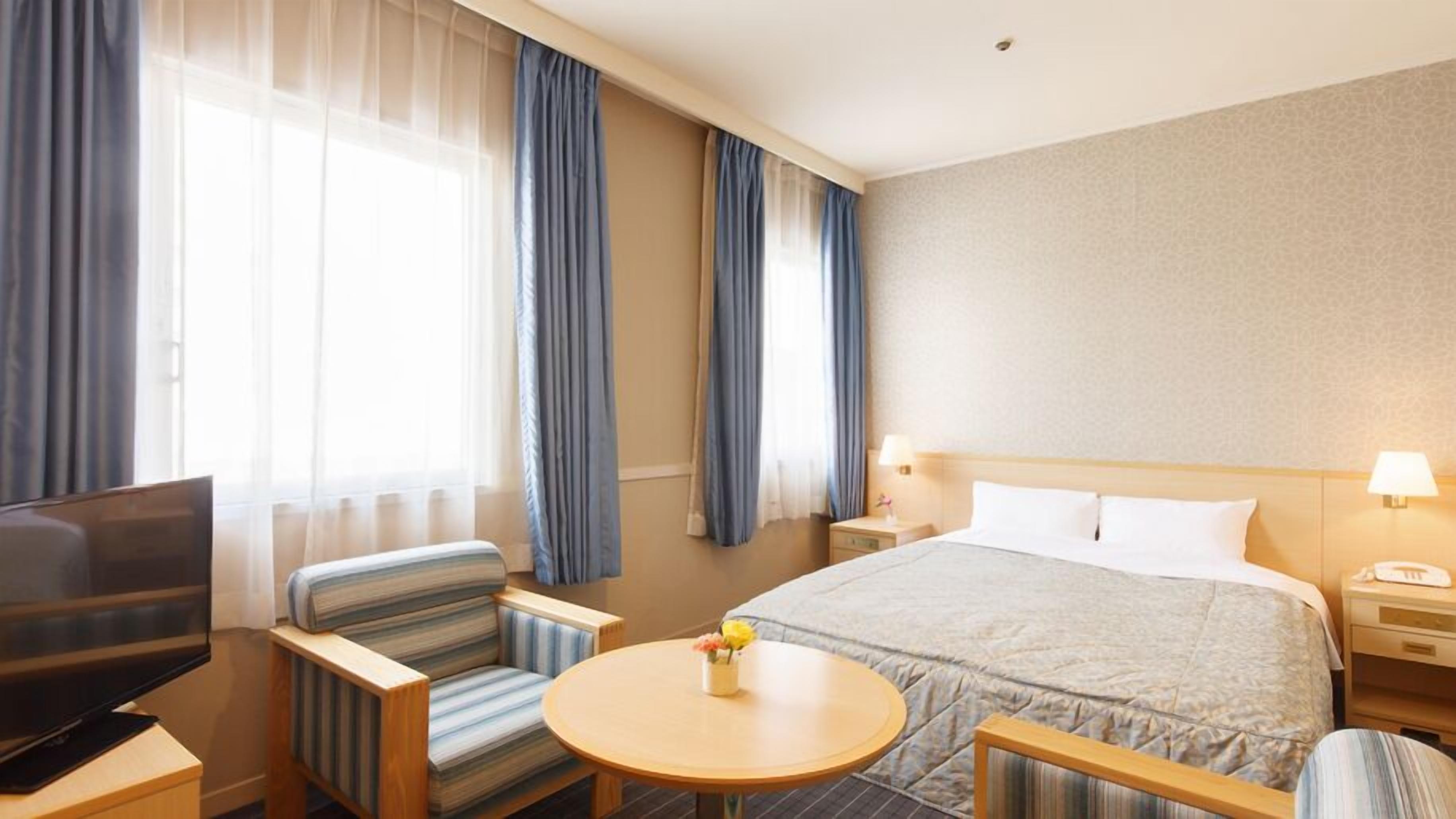 Quarto de hotel com uma cama, televisão, escrivaninha, cadeira e uma pequena mesa com flores.