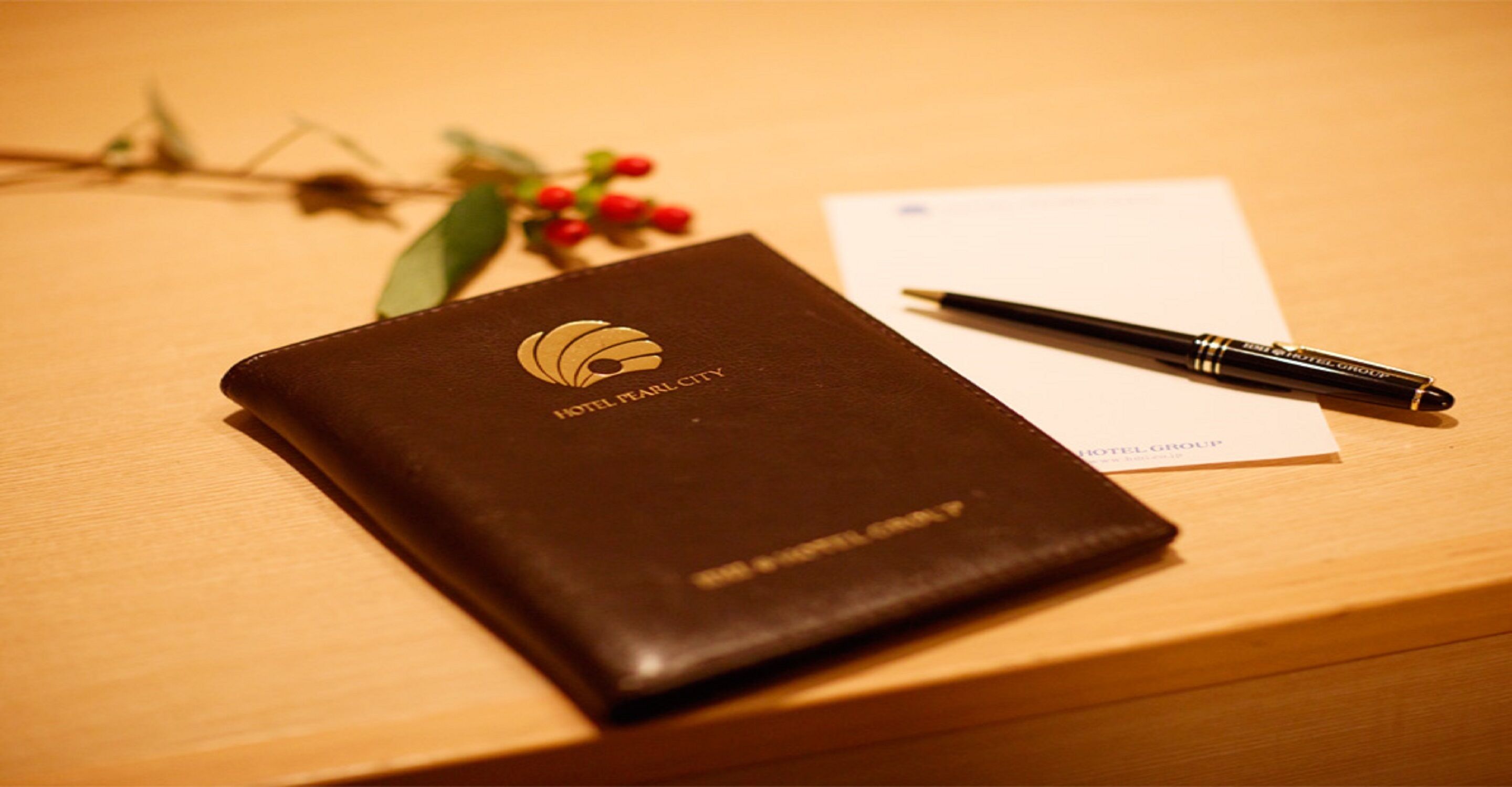 Ein braunes, ledergebundenes Buch mit dem aufprägem 'HOTEL PEARL CITY' auf dem Einband, auf einer Holzoberfläche zusammen mit einem schwarzen Stift und einigen Blättern.