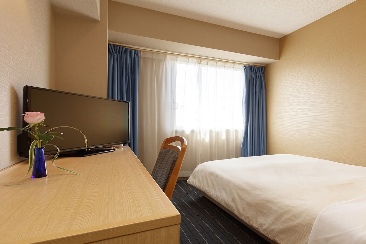 Ein Hotelzimmer mit Bett, Schreibtisch mit Fernseher, Stuhl und einem fensterbepflanzten Fenster.