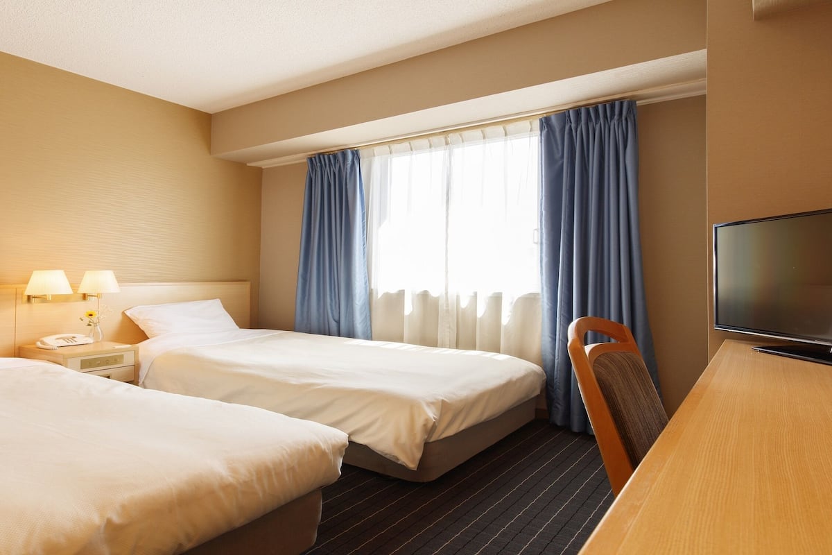 Ein Hotelzimmer mit zwei Betten, einem Schreibtisch, einem Fernseher und einem Fenster mit Vorhängen.
