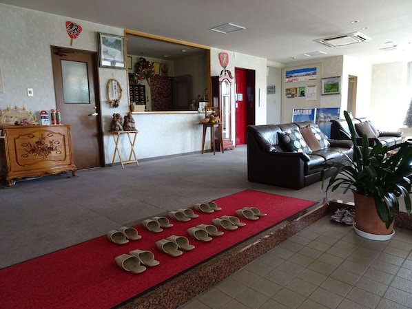 A hotel lobby with a reception desk, a sofa, a wooden cabinet, and a red carpet leading to a row of shoes.