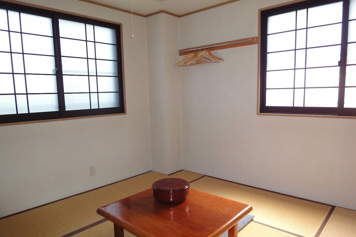 A room with tatami flooring, a wooden table with a dark wooden bowl, and two windows with black frames.
