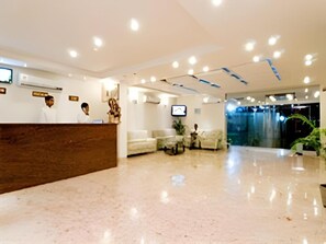 A hotel reception area with a wooden reception desk, a seating area with sofas, a television screen, and a water feature in the background. - Hotel Deviram Palace (Agra)