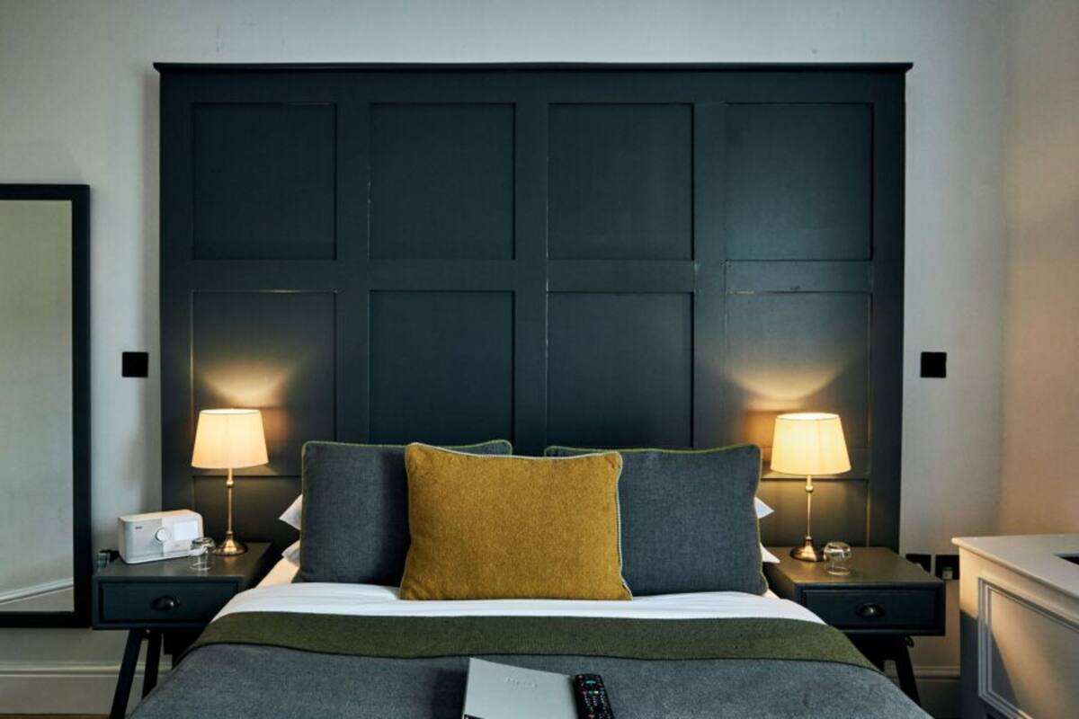 A hotel room with a large dark headboard, two bedside lamps, a bed with grey bedding, and a remote control on the bed.