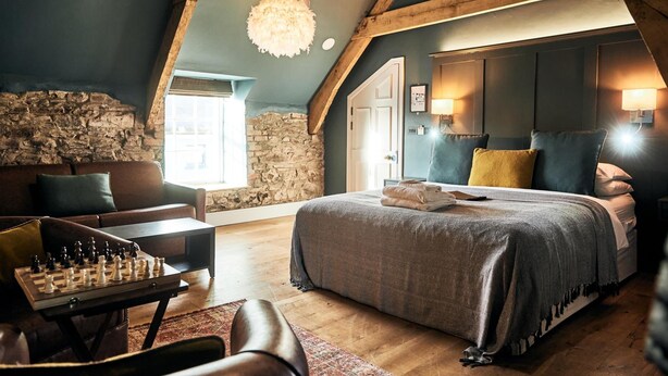 Um quarto aconchegante com parede de pedra, uma cama grande com almofadas, teto de madeira e um tabuleiro de xadrez sobre uma mesa.